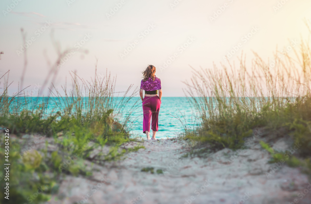 Rear view of a young woman walking on the beach towards the sea alone. Coastal summer evening time for relaxing, meditation, and contemplation. Rear view of a young woman walking on the beach towards the sea alone. Coastal summer evening time for relaxing, meditation, and contemplation.