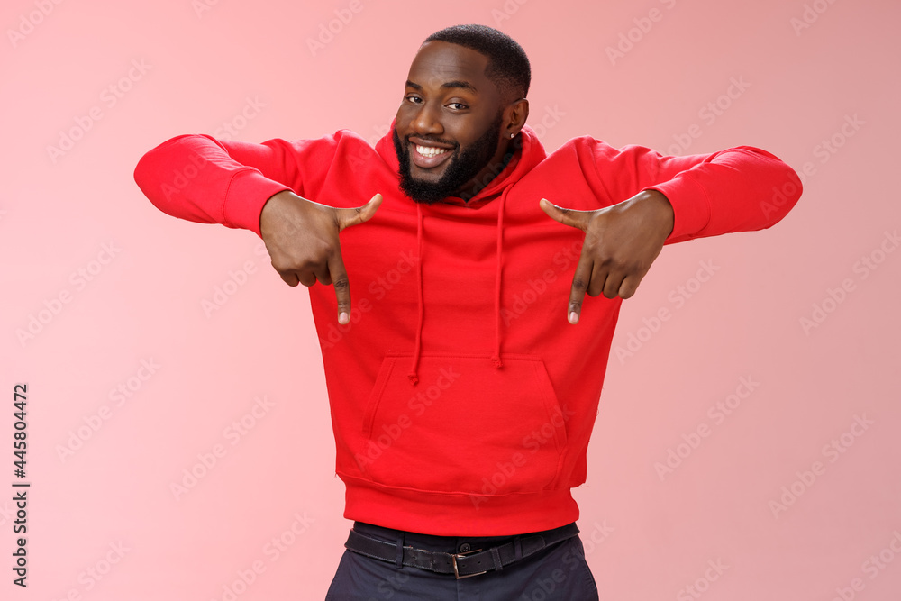 Sassy young charismatic handsome bearded black guy in red hoodie pointing down index fingers smiling broadly showing awesome copy space solution cool product for you standing pink background Stock Pho...