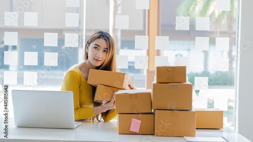 Independent Asian woman SME business owner online shopping working on laptop computer with parcel box on table at home - online SME business and online shipping and shopping concept.