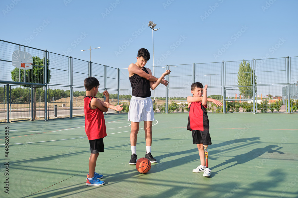 Basketball coach doing arm stretching exercises with two children, one of them has a leg prosthesis. Trainer training two kids.