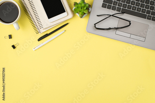 Top view photo of laptop glasses plant pens binder clips cup of drink and cellphone on two spiral reminders on isolated pastel yellow background with blank space