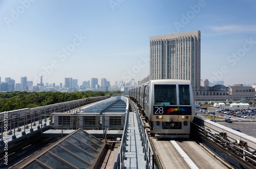Scenery of a train traveling on the elevated rail of Yurikamome Line passing by a modern building & a skyline of high-rise skyscrapers in background under blue clear sunny sky in Odaiba, Minato, Tokyo
