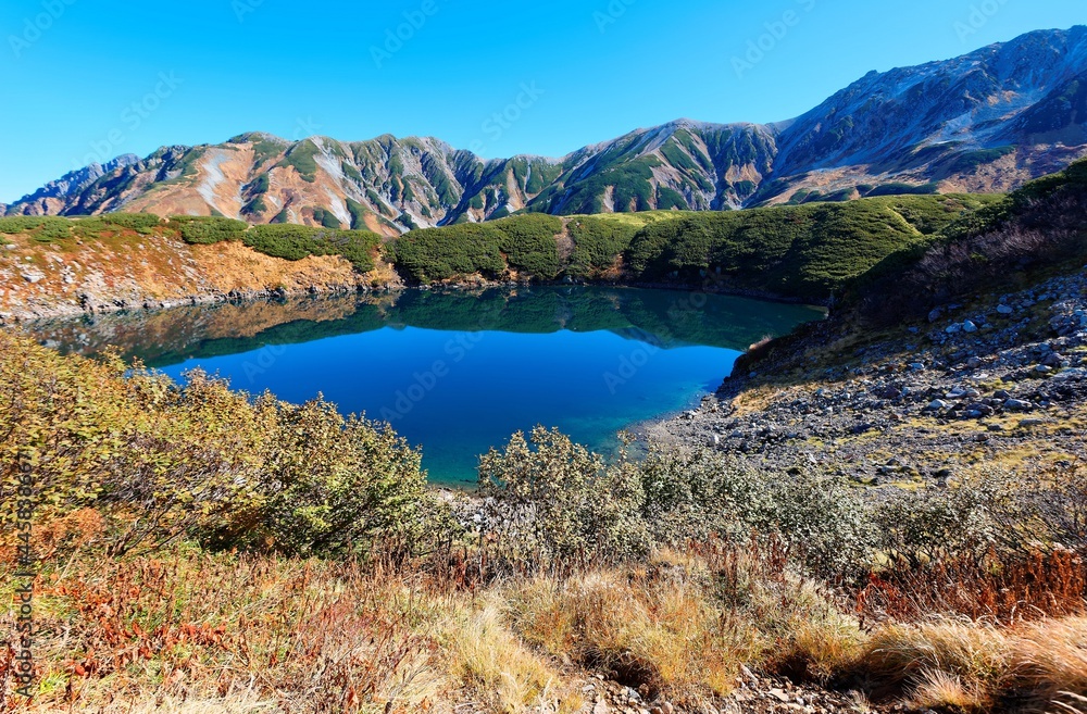 Fall scenery of beautiful Mikurigaike Pond, a crater lake in Murodo Tateyama, Toyama, Japan, with majestic mountain in background under clear blue sunny sky on a brisk autumn day ~A hiking destination