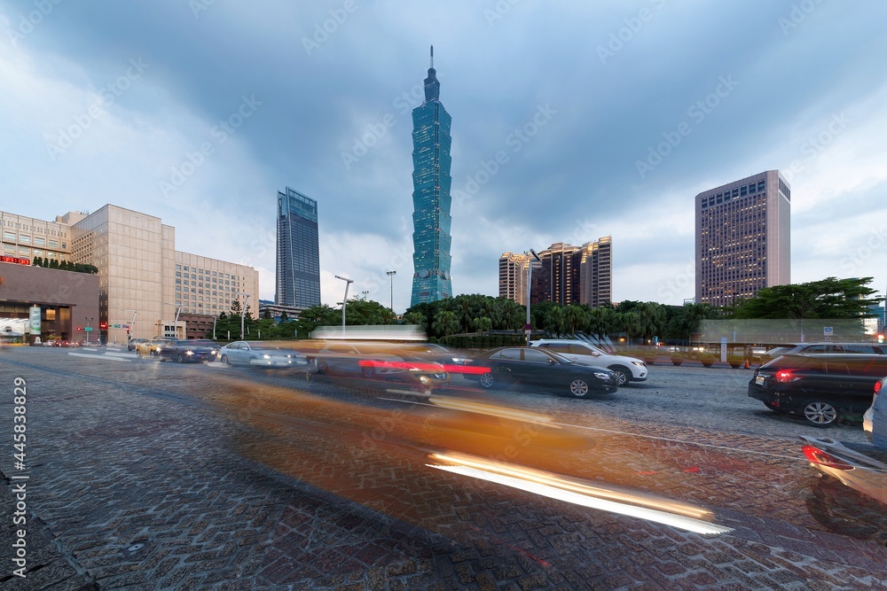 Naklejka premium Scenery of a busy street corner at rush hour in Downtown Taipei, capital city of Taiwan, with light trails of cars dashing by the City Hall & Taipei 101 Tower in Xinyi District under moody cloudy sky