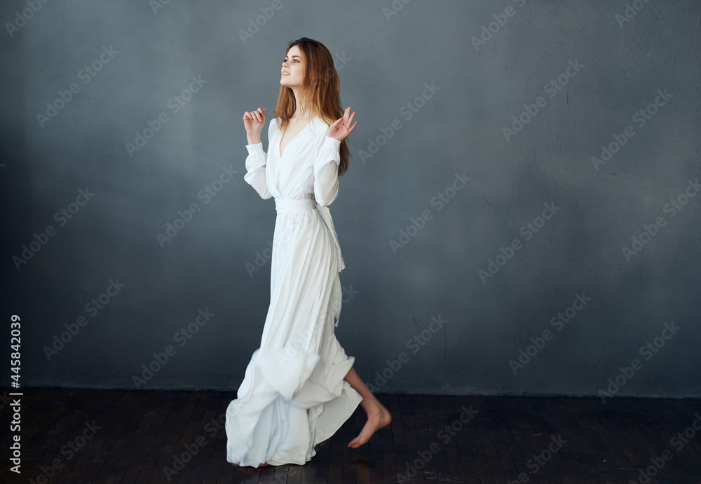 Woman in white dress posing on a dark background barefoot in full growth