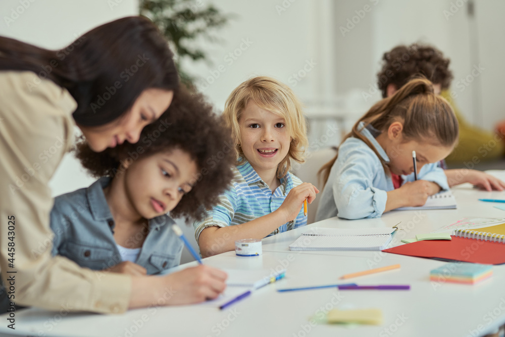 Making progress. Smiling little schoolboy looking at camera while sitting at the table in elementary school classroom together with other kids Making progress. Smiling little schoolboy looking at camera while sitting at the table in elementary school classroom together with other kids