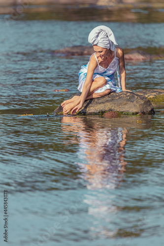 Fototapeta Naklejka Na Ścianę i Meble -  Young woman with a towel on her head at the sea. The lady touches the water with her hands. The concept of summer outdoor recreation, relaxation. good weather