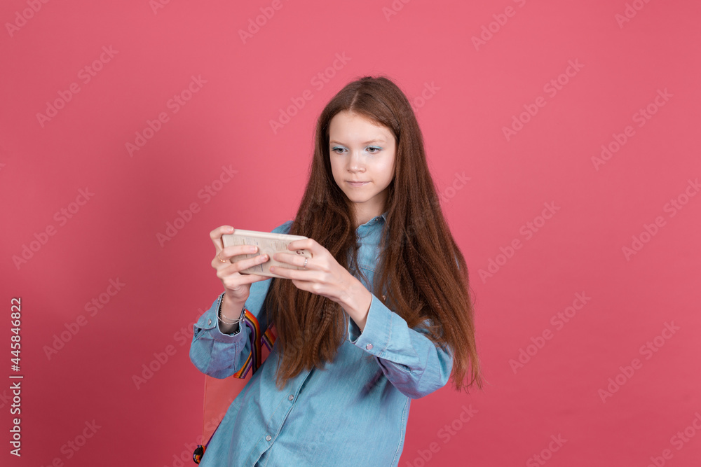 Little kid girl 13 years old in blue denim jacket isolated on red background schoolgirl with backpack with mobile phone playing games Little kid girl 13 years old in blue denim jacket isolated on red background schoolgirl with backpack with mobile phone playing games