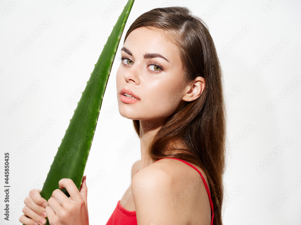 Naklejka premium beautiful woman with clean skin holds a green aloe leaf near her face on a light background