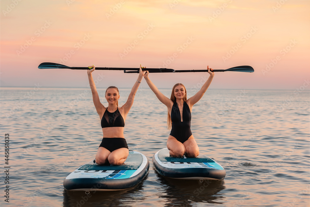 Surfing. Two happy women sitting on a sup board and holds up paddles ...