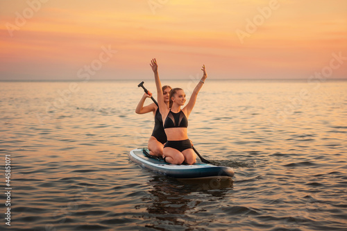 Summer surfing. Two happy women swimming on a sup board. In the background, the ocean and the sunset. Summer extreme recreation