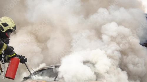 Professional firemen helping to extinguish with a fire extinguisher broken car after road accident in countryside. A lot of flames and smoke come out of the car. Firefighters save people Slow motion