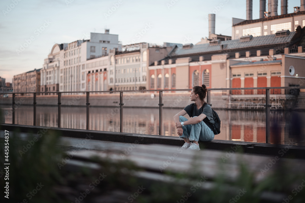 Young pensive beautiful woman sitting on the embankment in the city at sunrise