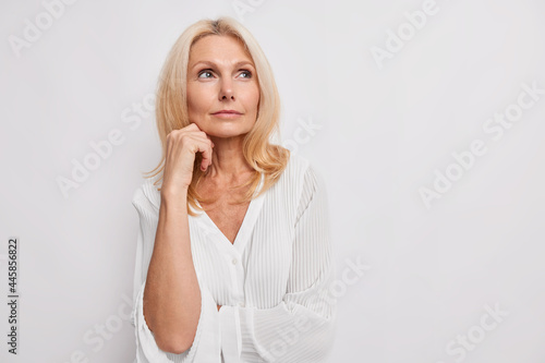Thoughtful blonde middle aged woman ponders on something keeps hand near face has healthy skin minimal makeup makes choice wears white blouse poses indoor blank copy space for your promotion