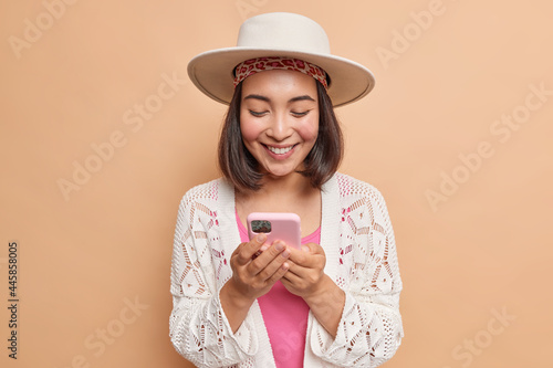 Happy Asian lady with dark hair wears hat and white coat looks gladfully at smartphone screen reads text message stands against beige background downloads new aplication. Technology concept.