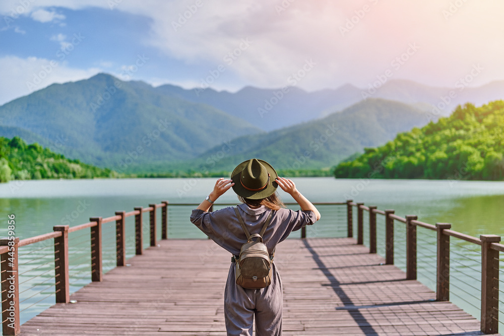 Obraz premium Traveler girl backpacker standing alone on pier and staring at lake and mountains. Enjoying beautiful freedom moment life and serene quiet peaceful atmosphere in nature. Back view