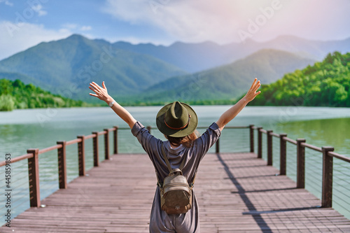 Tableau sur toile Traveler girl backpacker with open raised arms standing alone on pier and staring at lake and mountains