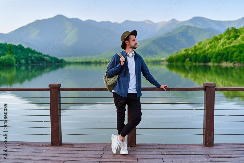 Fototapeta premium Hipster traveler backpacker standing alone on pier with lake and mountains view. Enjoying beautiful freedom moment life and serene quiet peaceful atmosphere in nature