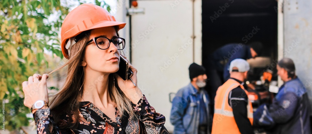 An engineer girl in a safety helmet talking on the phone against the background of construction and workers. Banner. An engineer girl in a safety helmet talking on the phone against the background of construction and workers. Banner.