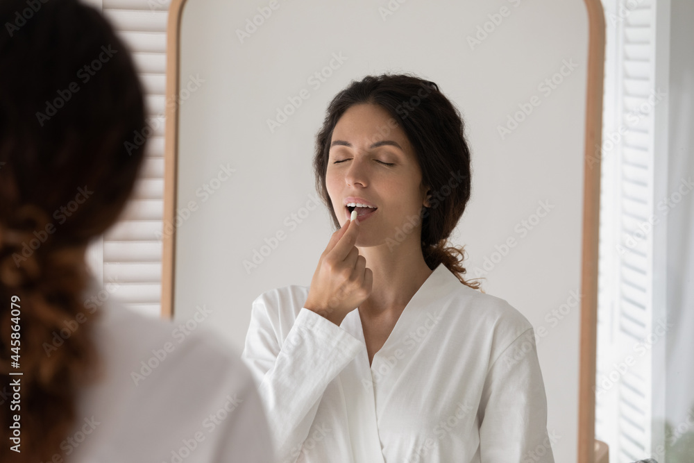 Taking meds. Young latin woman standing by mirror at bathroom drinking probiotic multivitamin tablet. Millennial female in morning gown swallowing oral contraceptive pill food supplement preparation