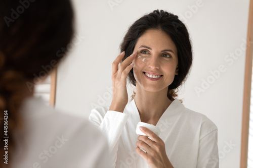 Fresh look every day. Smiling young hispanic woman apply nourishing cream to delicate face skin area under eyes. Beautiful millennial female put points of facial creme by mirror doing daily routine