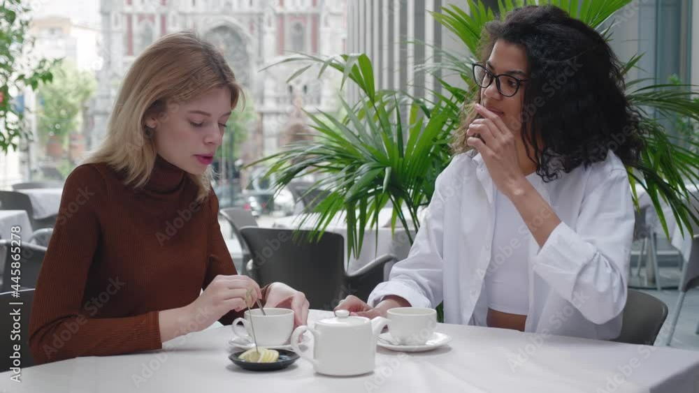 Two attractive girls sitting at the street cafe, communicate and sharing stories while, spending relax outdoor. Two diverse women friends are sitting in a cafe and talking Two attractive girls sitting at the street cafe, communicate and sharing stories while, spending relax outdoor. Two diverse women friends are sitting in a cafe and talking