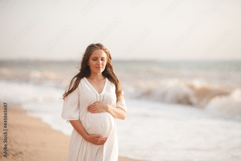 Smiling pregnant woman wear white dress hol tummy walk at beach over sea nature background outdoors. Summer vacation season. Motherhood. Maternity. Smiling pregnant woman wear white dress hol tummy walk at beach over sea nature background outdoors. Summer vacation season. Motherhood. Maternity.