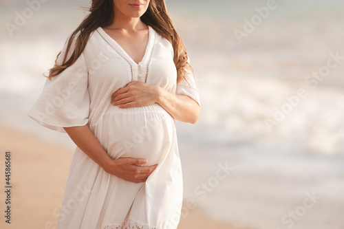 Smiling pregnant woman wear white dress hol tummy walk at beach over sea nature background outdoors. Summer vacation season. Motherhood. Maternity. #445865681