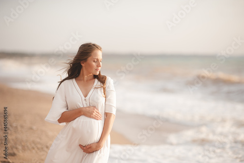 Canvas Print Smiling beautiful pregnant woman wear stylish white dress hold belly walk at beach over waves of sea outdoors