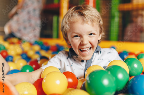 Happy little kid boy playing at balls pool playground. Happy kid playing in pool with plastic multicolored balls. Young boy smiling at the camera and having fun in indoor park playground