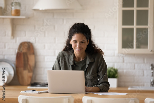 Young casual latin lady sit at kitchen table before laptop writing business email managing electronic documents. Busy millennial female expert freelancer working from home on startup project solution