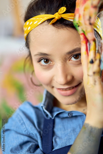 Close up portrait of happy young woman, female artist looking at camera, posing with her hands in paint while creating painting