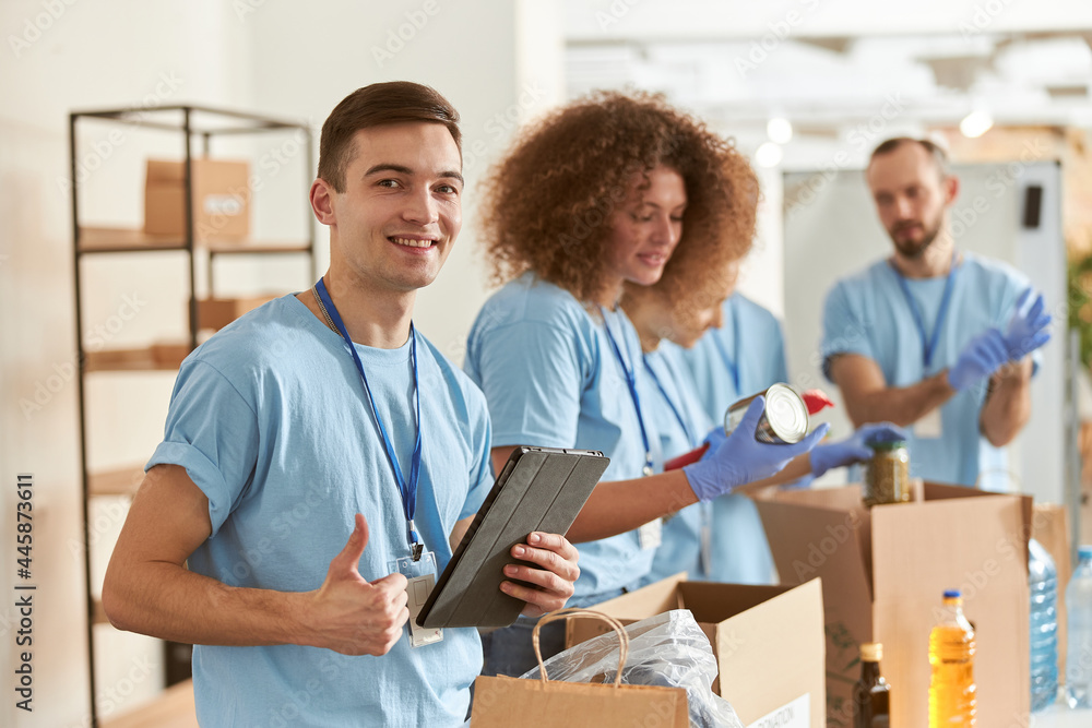 Portrait of happy male volunteer smiling at camera, showing thumbs up and holding tablet pc while sorting, packing food stuff in cardboard box, working on donation project with team. Focus on man Portrait of happy male volunteer smiling at camera, showing thumbs up and holding tablet pc while sorting, packing food stuff in cardboard box, working on donation project with team. Focus on man