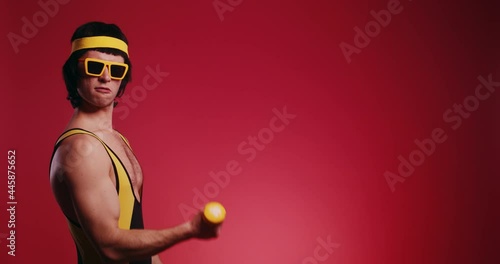 Funny retro handsome athlete man in yellow tracksuit and sunglasses from 80's with dumbbells makes the workout isolated on red background. Retro sport hipster show off and posing in front of camera.