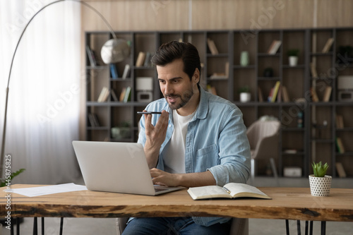 Fotografie Smart skilled happy young businessman sitting at table with computer in modern home office, recording audio message for client on smartphone or holding voicemail distant conversation with colleagues