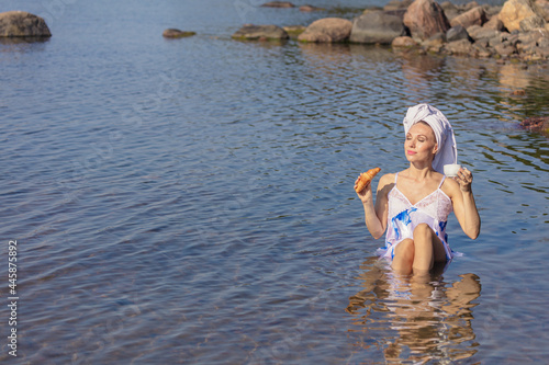 Young woman holding a cup of coffee and a croissant. a towel on your head. Lady sits in the sea. Concept of summer vacation  skin care and health  relaxation  spa  breakfast in nature
