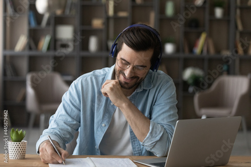 Happy motivated young 30s man in eyeglasses listening educational lecture in wireless headphones, writing notes in copybook, improving professional knowledge watching online webinar on computer.