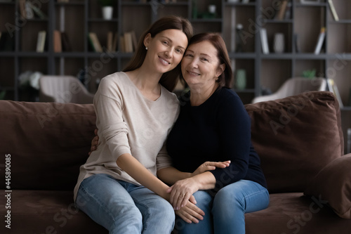 Happy grownup daughter and mature 60s mother resting on couch at home, hugging, looking at camera, smiling, enjoying leisure time together. Intergenerational family relations. Head shot portrait