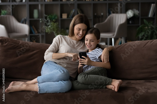 Happy kid and mom relaxing on sofa at home, using app on mobile phone, making video call, playing online game, shopping on internet together, browsing social media. Mother and girl looking at screen
