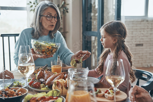 Beautiful senior woman putting salad for little girl while having dinner together