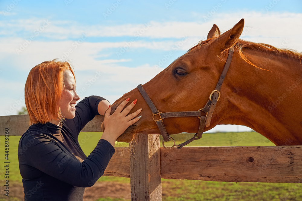 Obraz premium Red-haired girl stroking horse in paddock on a summer sunny day