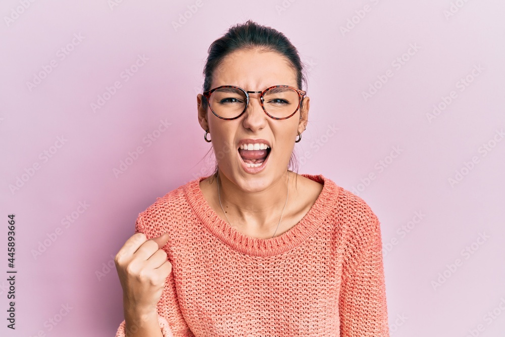 Young caucasian woman wearing casual clothes and glasses angry and mad raising fist frustrated and furious while shouting with anger. rage and aggressive concept. Young caucasian woman wearing casual clothes and glasses angry and mad raising fist frustrated and furious while shouting with anger. rage and aggressive concept.