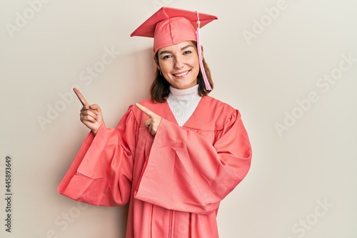 Young caucasian woman wearing graduation cap and ceremony robe smiling and looking at the camera pointing with two hands and fingers to the side.
