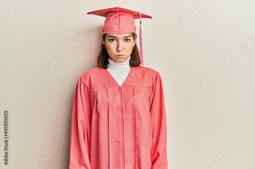 Young caucasian woman wearing graduation cap and ceremony robe puffing cheeks with funny face. mouth inflated with air, crazy expression.