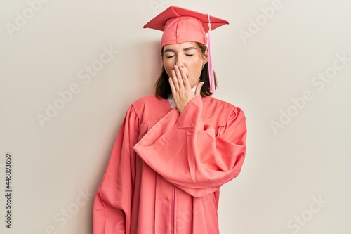 Young caucasian woman wearing graduation cap and ceremony robe bored yawning tired covering mouth with hand. restless and sleepiness.