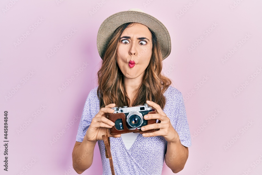 Young hispanic girl wearing summer hat holding vintage camera making fish face with mouth and squinting eyes, crazy and comical.