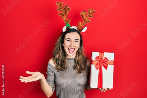 Young hispanic girl wearing deer christmas hat holding gift celebrating achievement with happy smile and winner expression with raised hand
