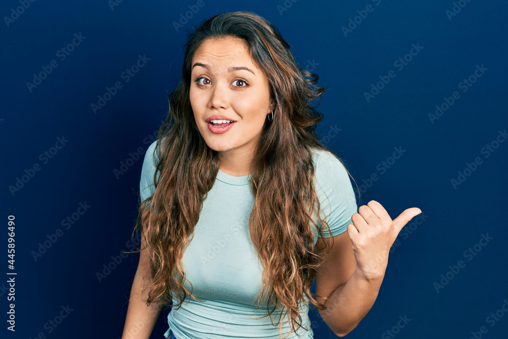 Young hispanic girl wearing casual clothes smiling with happy face looking and pointing to the side with thumb up. Young hispanic girl wearing casual clothes smiling with happy face looking and pointing to the side with thumb up.