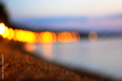 Blurred evening promenade by the sea in defocus. Yellow lantern lights on lampposts and blue calm sea at dusk. Night descends on a city. A strong blur. Beautiful seascape abstract background.