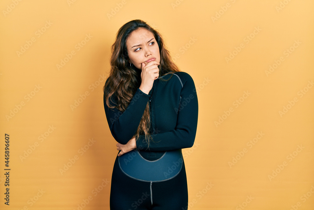 Young hispanic girl wearing diver neoprene uniform with hand on chin thinking about question, pensive expression. smiling with thoughtful face. doubt concept. Young hispanic girl wearing diver neoprene uniform with hand on chin thinking about question, pensive expression. smiling with thoughtful face. doubt concept.